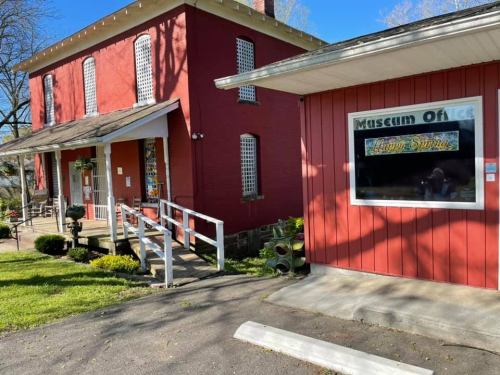 A red building with a porch and a sign for "Museum Offices" in front, surrounded by greenery and a clear blue sky.