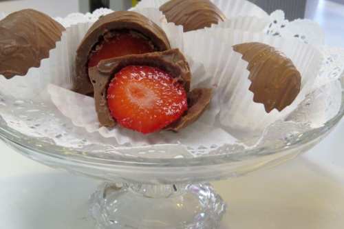 A glass dish holds chocolate-covered strawberries, some whole and one cut in half, revealing the juicy fruit inside.
