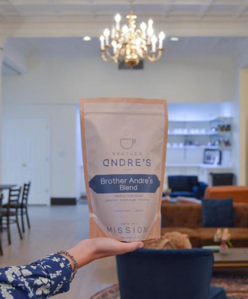 A hand holds a bag of Brother Andre's Blend coffee in a stylish room with a chandelier and cozy seating.