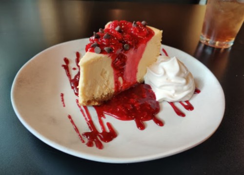 A slice of cheesecake topped with raspberry sauce, chocolate chips, and a dollop of whipped cream on a white plate.