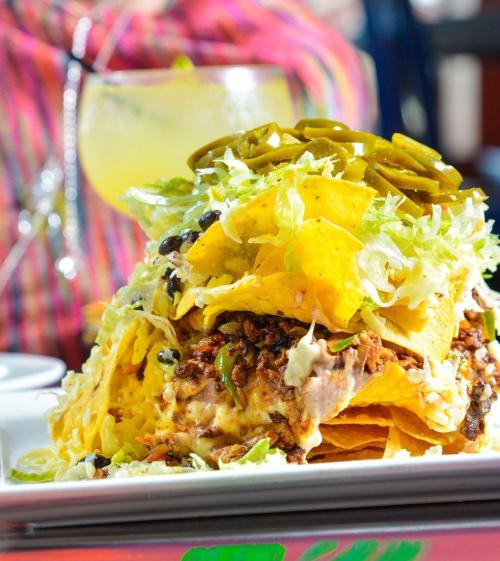 A colorful plate of nachos topped with shredded meat, lettuce, jalapeños, and cheese, served with a drink in the background.