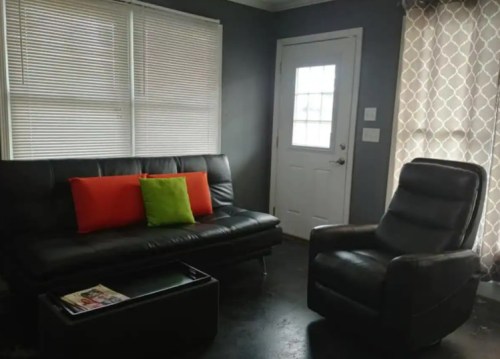 A cozy living room with a black sofa, colorful cushions, a recliner, and a door with a window.