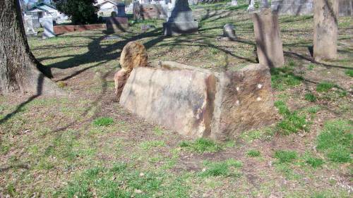 A weathered, large stone grave marker lies on the ground in a grassy cemetery, surrounded by trees and other gravestones.