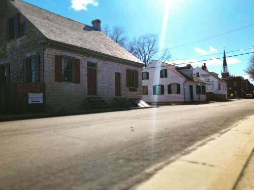 A quiet street lined with historic houses under a bright blue sky, featuring a church steeple in the background.