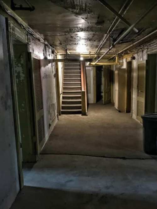 Dimly lit hallway with stairs at the end, featuring peeling walls and closed doors on either side.
