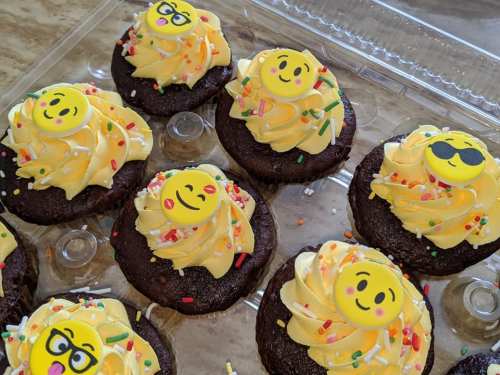 A tray of chocolate cupcakes topped with yellow frosting, sprinkles, and smiley face decorations.