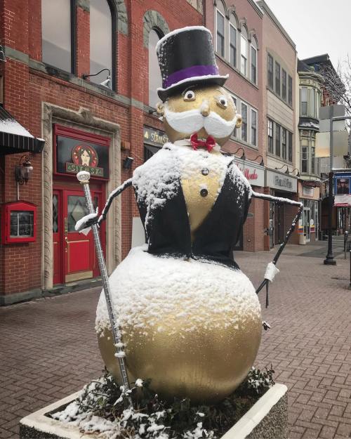 A large snowman with a top hat and monocle, covered in snow, stands in a quaint town square.