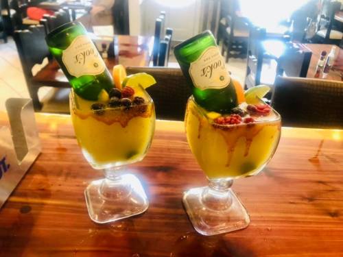 Two colorful cocktails with fruit garnishes and beer bottles nestled in the glasses, set on a wooden table.