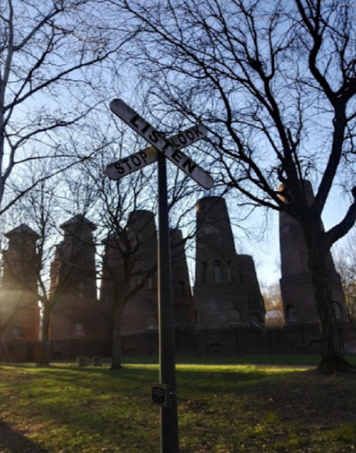 A weathered railroad sign stands in front of old, towering brick ruins, surrounded by bare trees and soft sunlight.