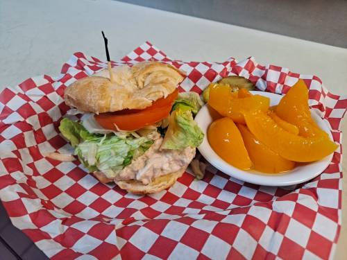 A croissant sandwich with lettuce, tomato, and chicken salad, served with a side of peach slices on a checkered paper.