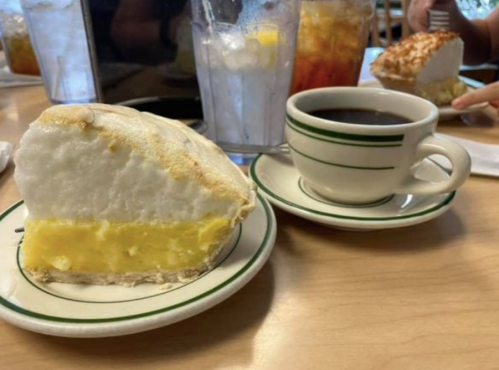 A slice of lemon meringue pie on a plate next to a cup of coffee on a wooden table.
