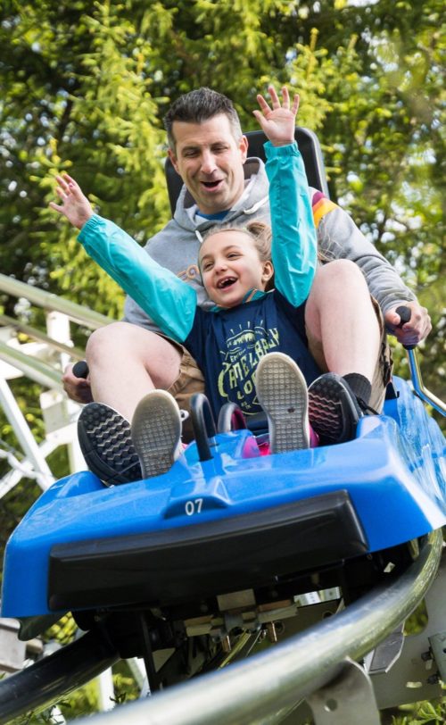 A man and a girl joyfully ride a roller coaster, both raising their hands in excitement as they descend.