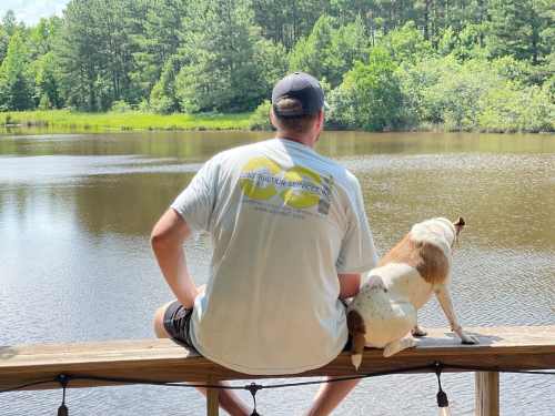 A person and a dog sit on a dock by a calm lake, surrounded by trees and greenery.
