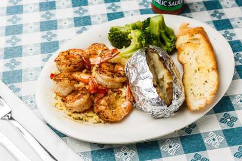 A plate with grilled shrimp, broccoli, a baked potato in foil, and garlic bread on a checkered tablecloth.