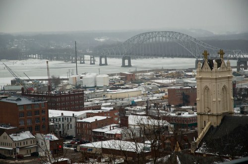 A snowy cityscape with a river and bridge in the background, featuring buildings and a church steeple in the foreground.