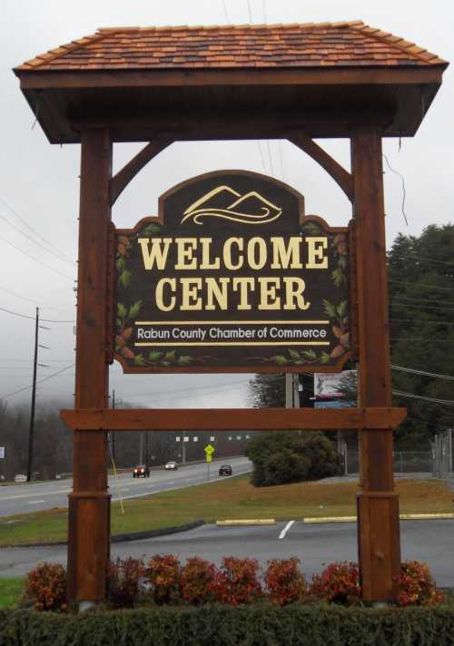 Sign for the Rabun County Welcome Center, featuring a wooden structure and mountain design, with a cloudy sky in the background.