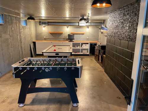 A spacious game room featuring a foosball table, a white game table, and modern lighting against a concrete wall.