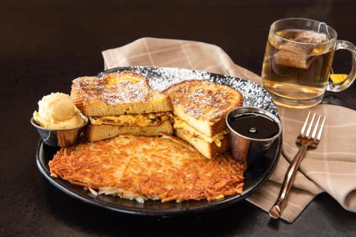 A plate of French toast with eggs, hash browns, butter, syrup, and a cup of tea on a dark table.