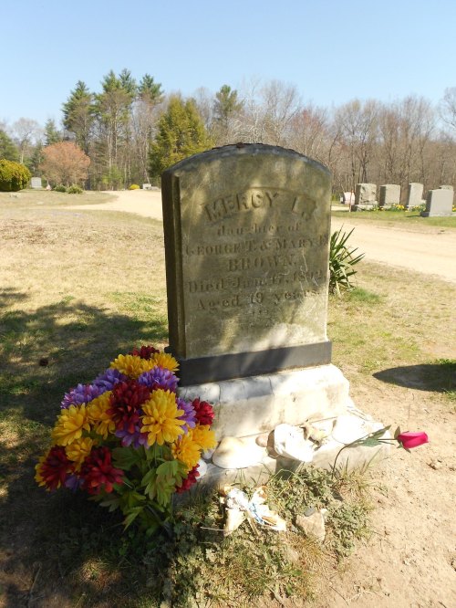 A weathered gravestone with the name "Mercy L. Brown," surrounded by colorful flowers in a cemetery setting.