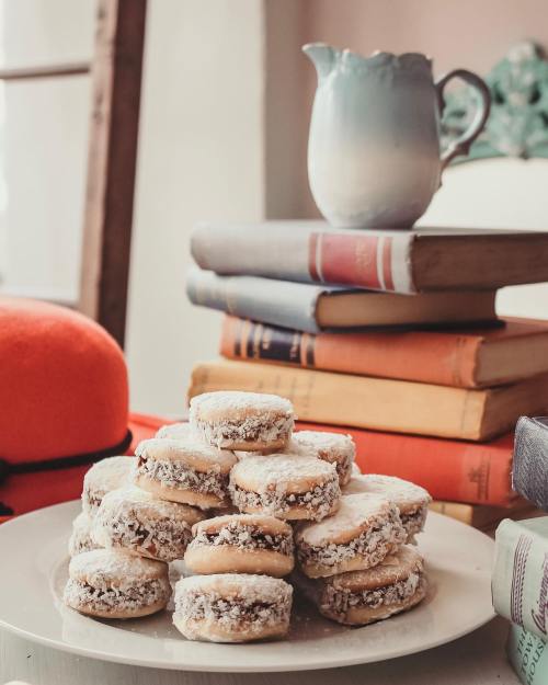 A plate of powdered sugar-dusted cookies stacked on a table with books and a small pitcher in the background.