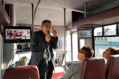 A man in a striped suit speaks animatedly on a bus, while passengers listen attentively. Screens show images in the background.