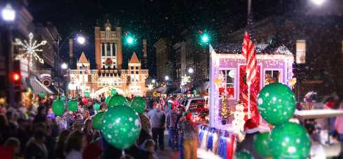 A festive night scene of a holiday parade with green balloons, lights, and a decorated float amidst a crowd.