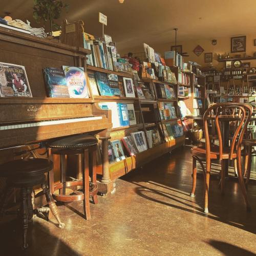 A cozy bookstore interior featuring a piano, wooden shelves filled with books, and a few chairs in warm sunlight.