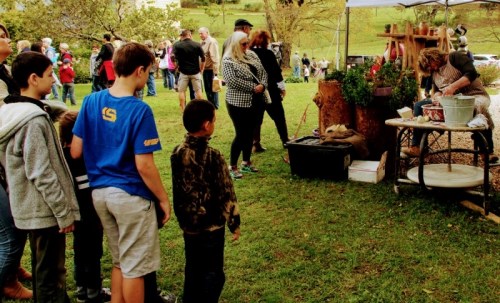 A group of children watch a demonstration at an outdoor event, with adults observing nearby.