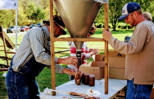 Two men in a park are filling jars with a funnel, surrounded by boxes and a table with food.