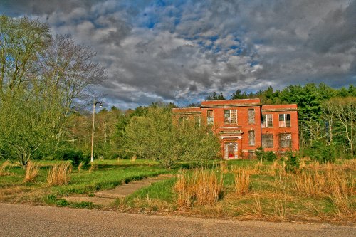 Abandoned brick building surrounded by overgrown grass and trees under a cloudy sky.