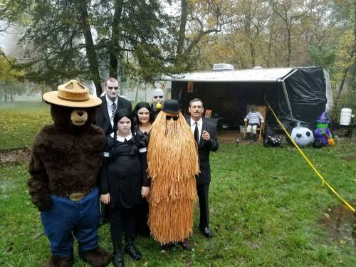 A group in costumes, including a bear, a ghost, and a figure with long hair, poses in front of a trailer in a foggy park.