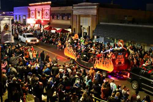 A festive parade scene with a decorated float, crowds of people, and buildings lit up at night.