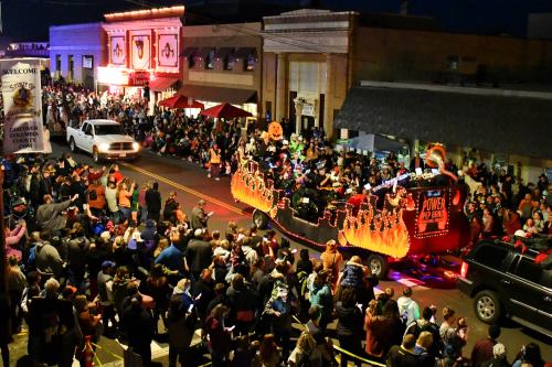 A festive parade scene with a decorated float, crowds of people, and buildings lit up at night.