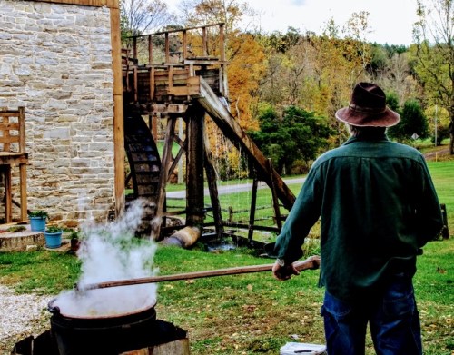 A person in a hat stirs a pot with steam, near a watermill and autumn trees in the background.
