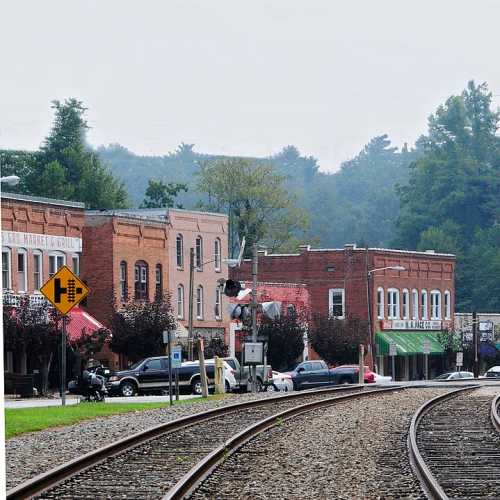 A quiet street scene with railroad tracks, historic buildings, and trees in the background on an overcast day.