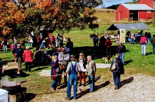 A lively outdoor gathering at a farm with people socializing, colorful trees, and red barns in the background.