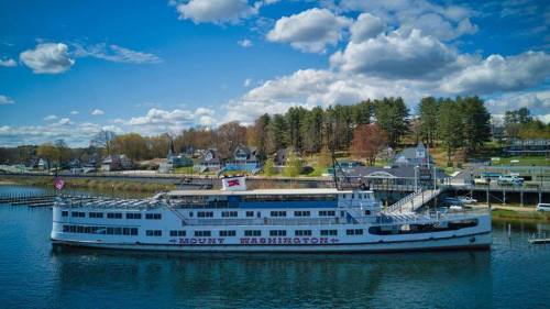 A large white boat named "Mount Washington" docked in a calm lake, surrounded by trees and houses under a blue sky.
