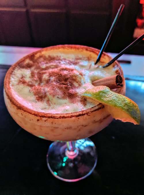 A frothy cocktail in a glass, garnished with a lime wedge and cinnamon stick, served on a bar with colorful lighting.