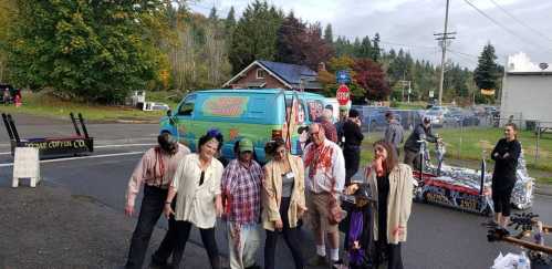 A group of people dressed as zombies pose on a street, with a colorful van and Halloween decorations in the background.