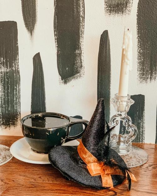 A black cup on a saucer beside a small witch hat and a candle in a glass holder against a painted wall.