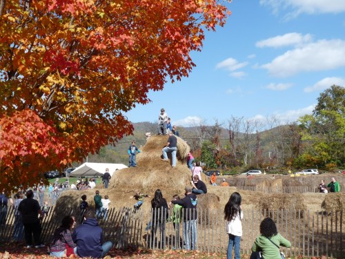Children play on a large haystack in a vibrant autumn setting with colorful trees and a clear blue sky.