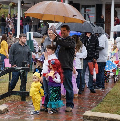 A group of people in costumes holding umbrellas during a rainy parade, with children in colorful outfits in the foreground.