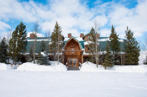 A large wooden lodge surrounded by snow and evergreen trees under a blue sky with clouds.