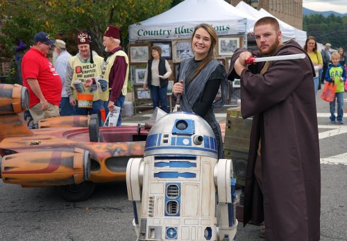 A woman poses with a large R2-D2 replica while a man in a Jedi costume stands beside her at a festival.
