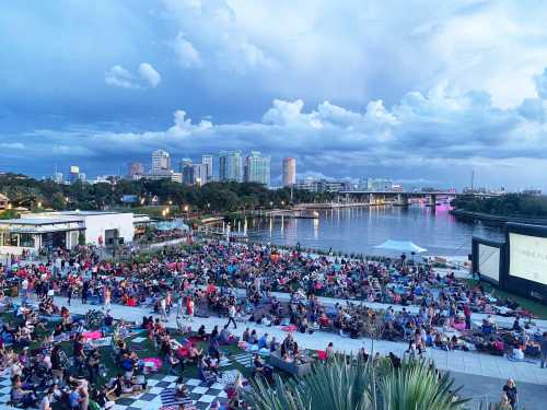 A large crowd gathers on a grassy area by a river, enjoying an outdoor movie with a city skyline in the background.