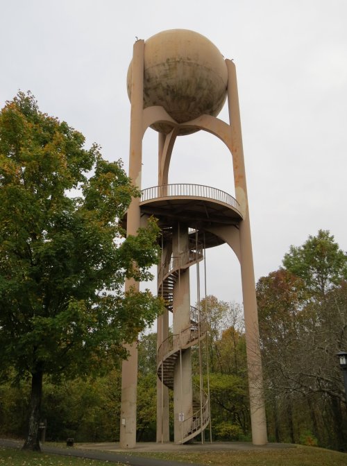 A tall water tower with a spherical top and a spiral staircase, surrounded by trees in a cloudy setting.