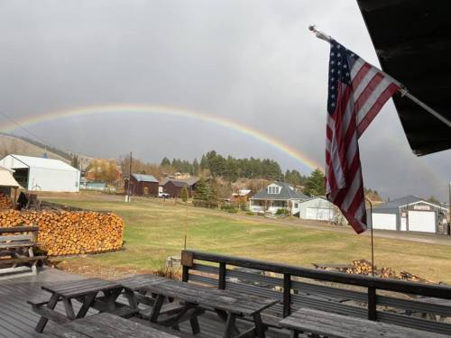 A rainbow arcs over a rural landscape with a flag waving in the foreground and wooden logs stacked nearby.