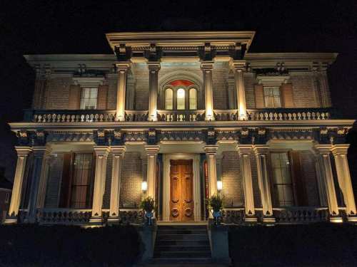 A grand, illuminated brick mansion at night, featuring ornate architectural details and a large wooden front door.