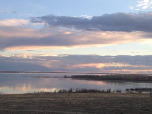 A serene landscape featuring a calm lake reflecting colorful clouds at sunset, with distant trees along the shore.