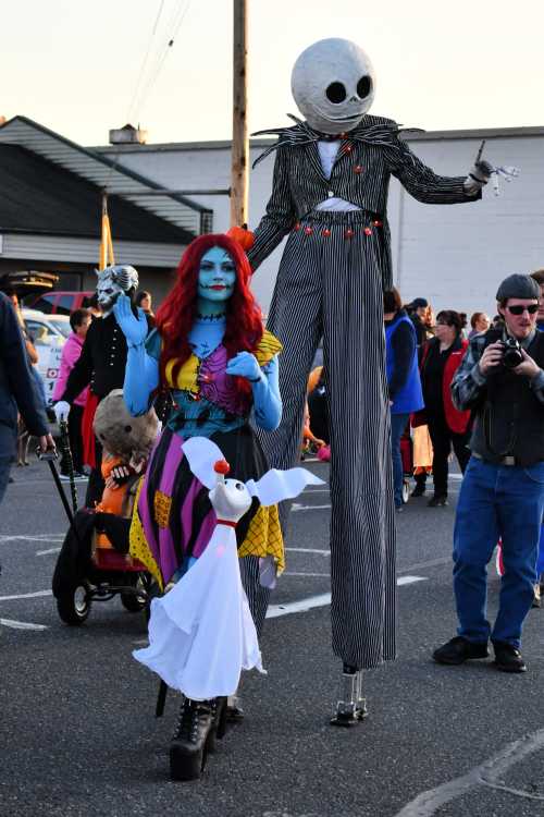 A tall figure in a striped suit and a skeleton head walks alongside a woman with blue skin and red hair, both in costumes.